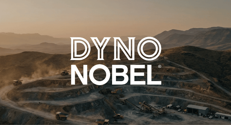Dyno Nobel logo overlaid on an aerial photograph of an active open-pit mining site at dusk, with haul trucks operating amid terraced quarry walls and mountains in the background.