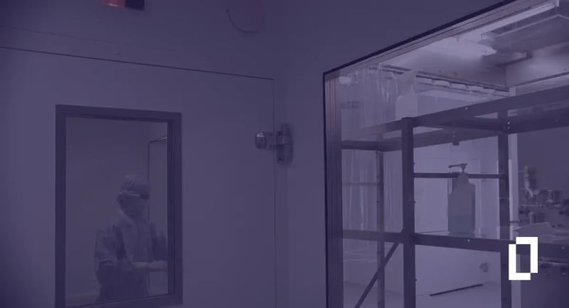 Pharmaceutical carbon accounting standards: clean room technician in protective clothing behind a glass window
