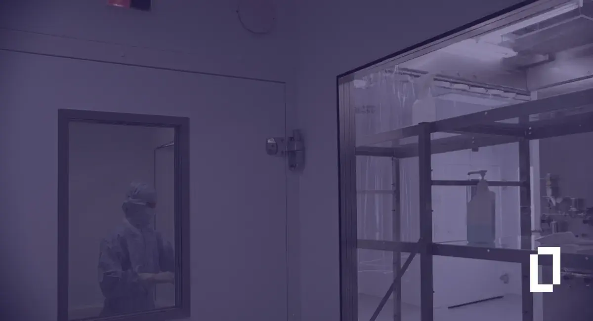 Pharmaceutical carbon accounting standards: clean room technician in protective clothing behind a glass window